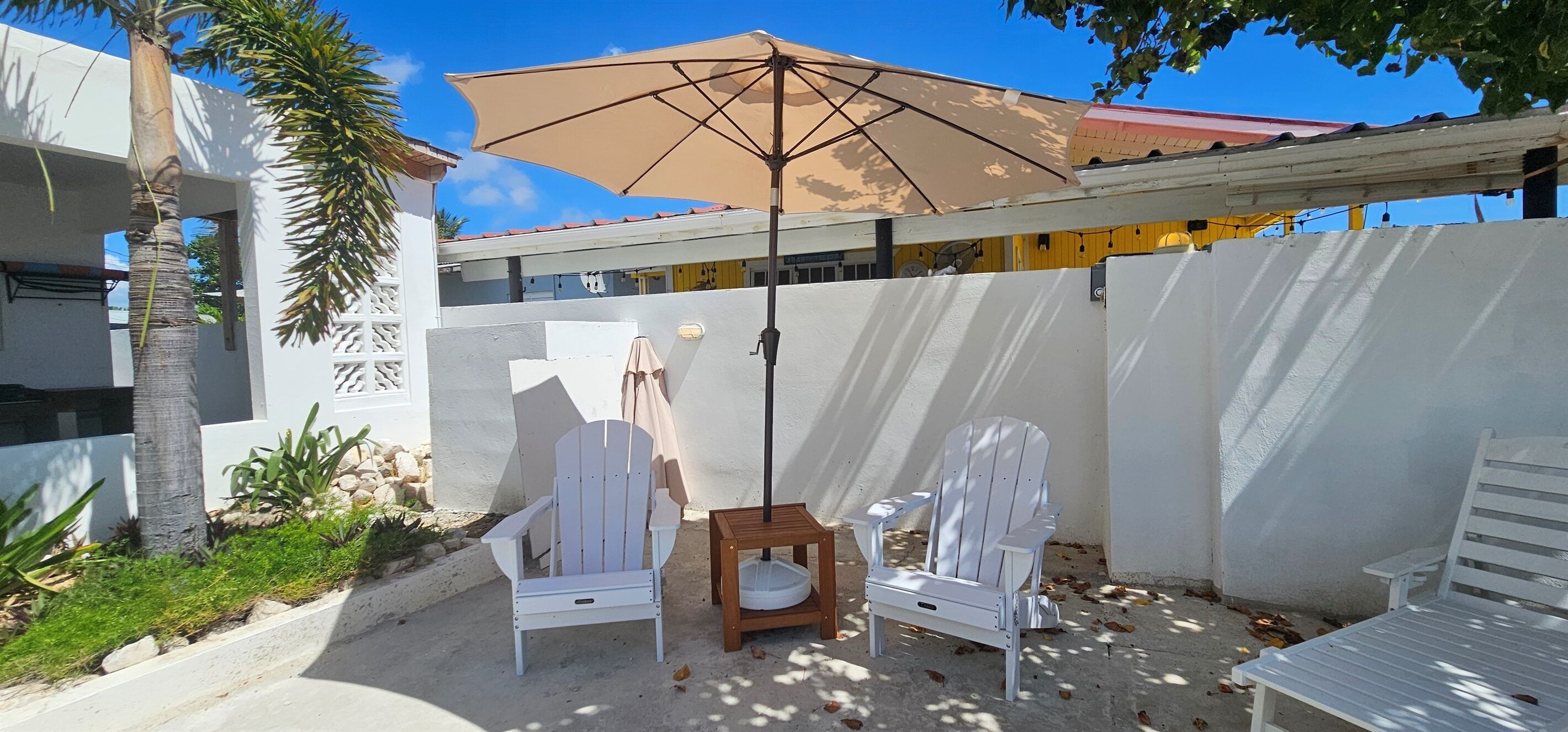 A patio area with two white Adirondack chairs, a wooden table, and a beige umbrella providing shade.