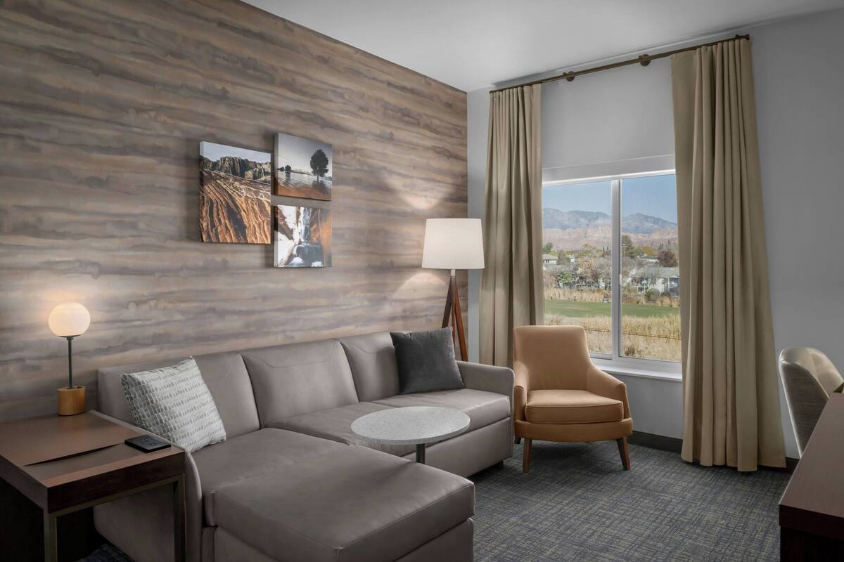 Una habitación de hotel moderna con un sofá modular, una mesa de centro redonda, un sillón individual, una lámpara y un ventanal con vistas a las montañas.