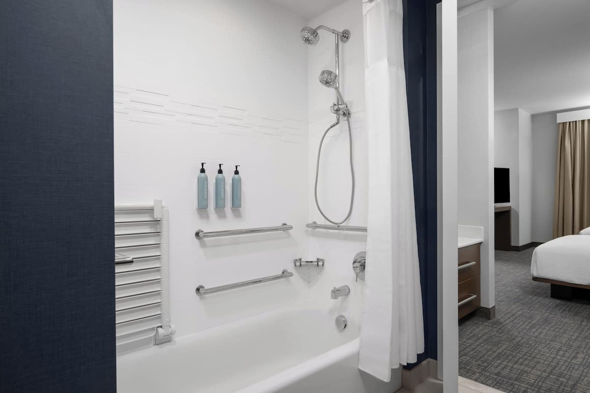 Un baño moderno con bañera, ducha y barras de apoyo. Hay artículos de aseo en la pared y una cama en la habitación contigua.