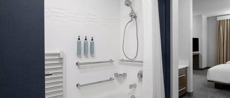 A modern bathroom with a bathtub, shower, and handrails. There are toiletries on the wall and a bed in the adjacent room.