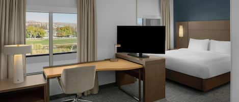 A hotel room with a desk, a bed, a TV, and a view of a landscape.