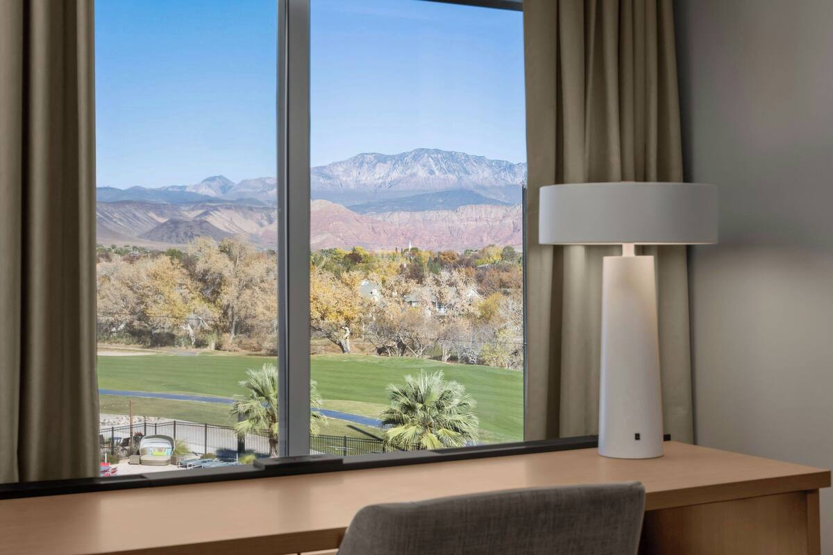 Habitación con un ventanal amplio que ofrece vistas a montañas y un parque. Hay una lámpara sobre un escritorio.