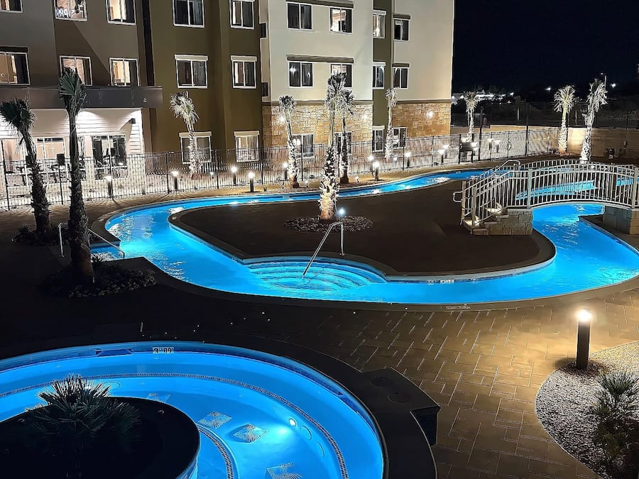 A nighttime view of a hotel pool area with multiple swimming pools, palm trees, and a multi-story building in the background.