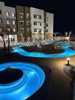Vista nocturna de un área de piscina de hotel con varias piscinas, palmeras y un edificio de varios pisos al fondo.