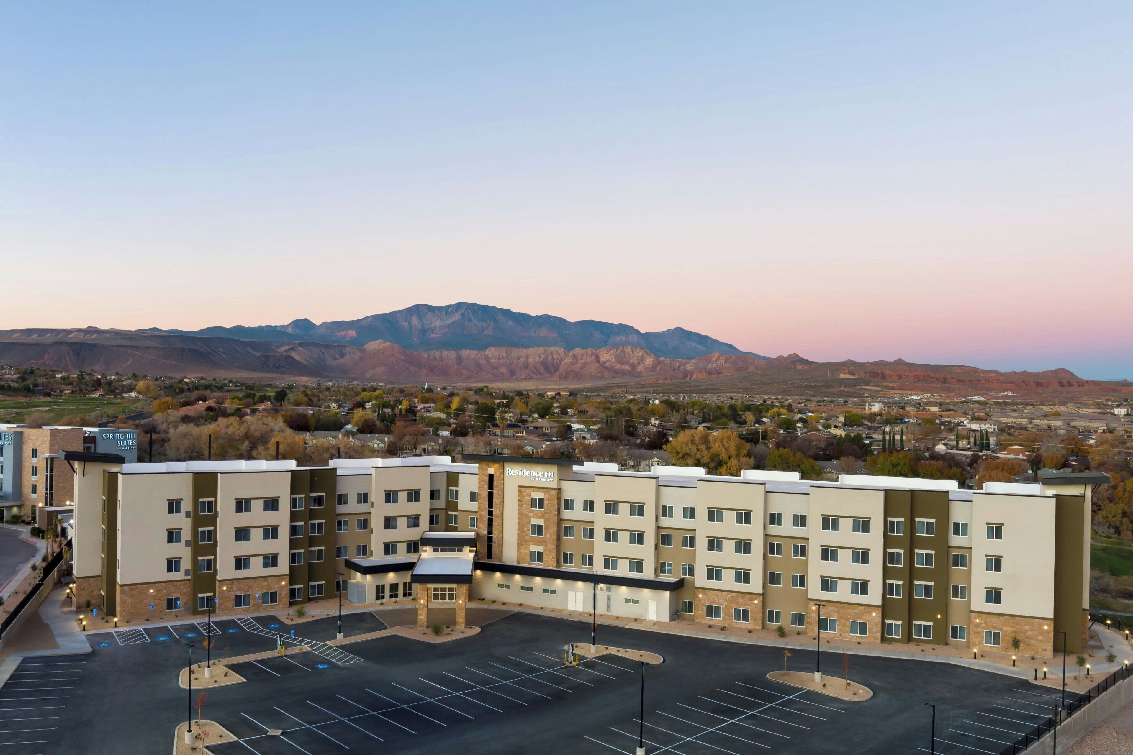 A multi-story hotel with a central courtyard, parking lot, and a mountain backdrop.