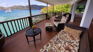 A balcony with a sofa, chairs, and a small table with flowers. It overlooks a body of water with boats and mountains in the background. - Hotel and restaurant in Bequia (Bequia)