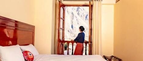 Una cama con estructura de madera, cubierta blanca y almohadas, techo de madera y vista a un paisaje montañoso a través de una puerta.