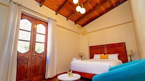 Habitación de hotel con techo de madera, cama con ropa blanca, una mesa redonda y un objeto dorado, y una silla azul.