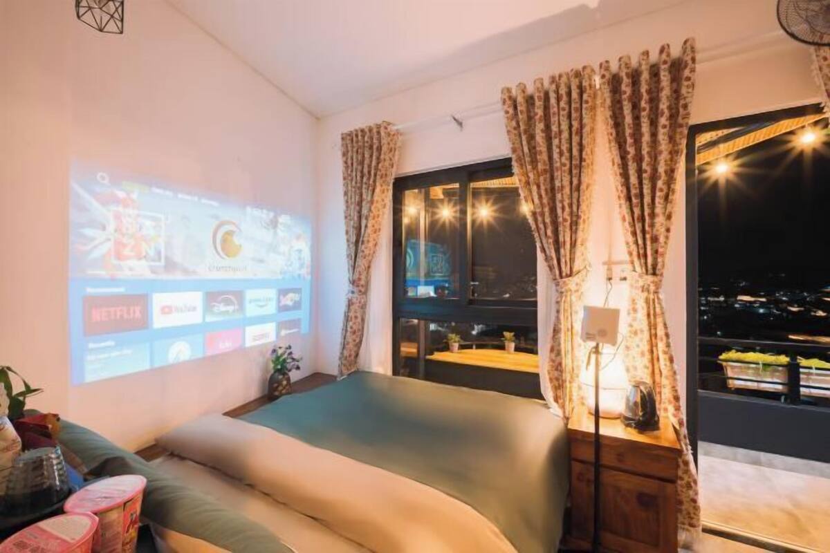 A bedroom with a bed, curtains, a projector screen, and a view of the city at night.