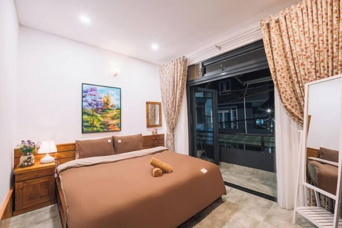 A bedroom with a large bed, a nightstand, a lamp, a painting, and a view of a cityscape through a window.