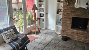 A cozy living room with a brick fireplace, a large window with a view of the garden, a comfortable sofa with a patterned cushion, and a small wooden side table with a lamp.