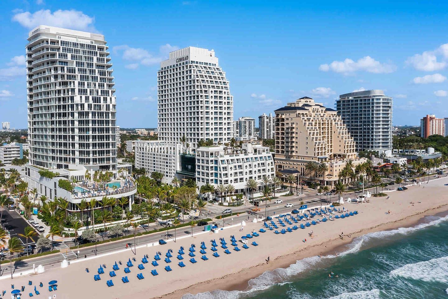 A coastal cityscape with high-rise hotels, a beach with rows of blue lounge chairs, and a clear blue sky.