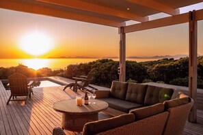 Eine Holzterrasse mit Gartenmöbeln, einem Pool und Blick auf den Sonnenuntergang über dem Meer.