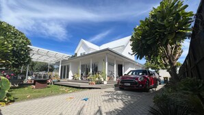 A white house with a covered porch, a red car parked in front, and various potted plants around.