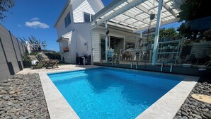 A rectangular swimming pool with a stone border, surrounded by a patio area with chairs and a table.