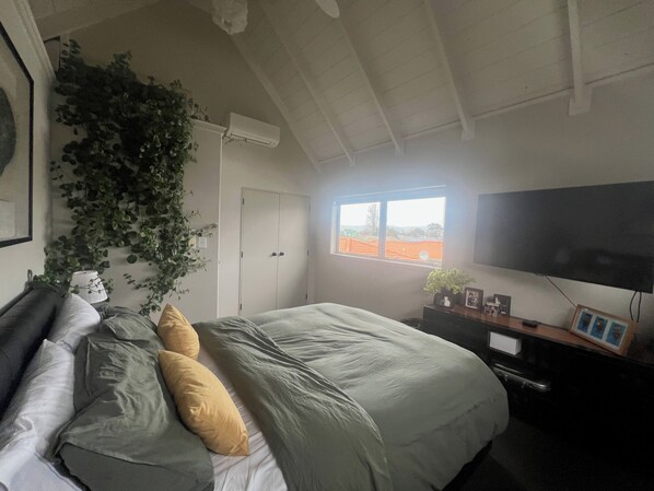 A bedroom with a bed, a TV, a wardrobe, and a window with a view of a red-roofed building. - Papamoa beach house with pool and indoor outdoor living (Tauranga)