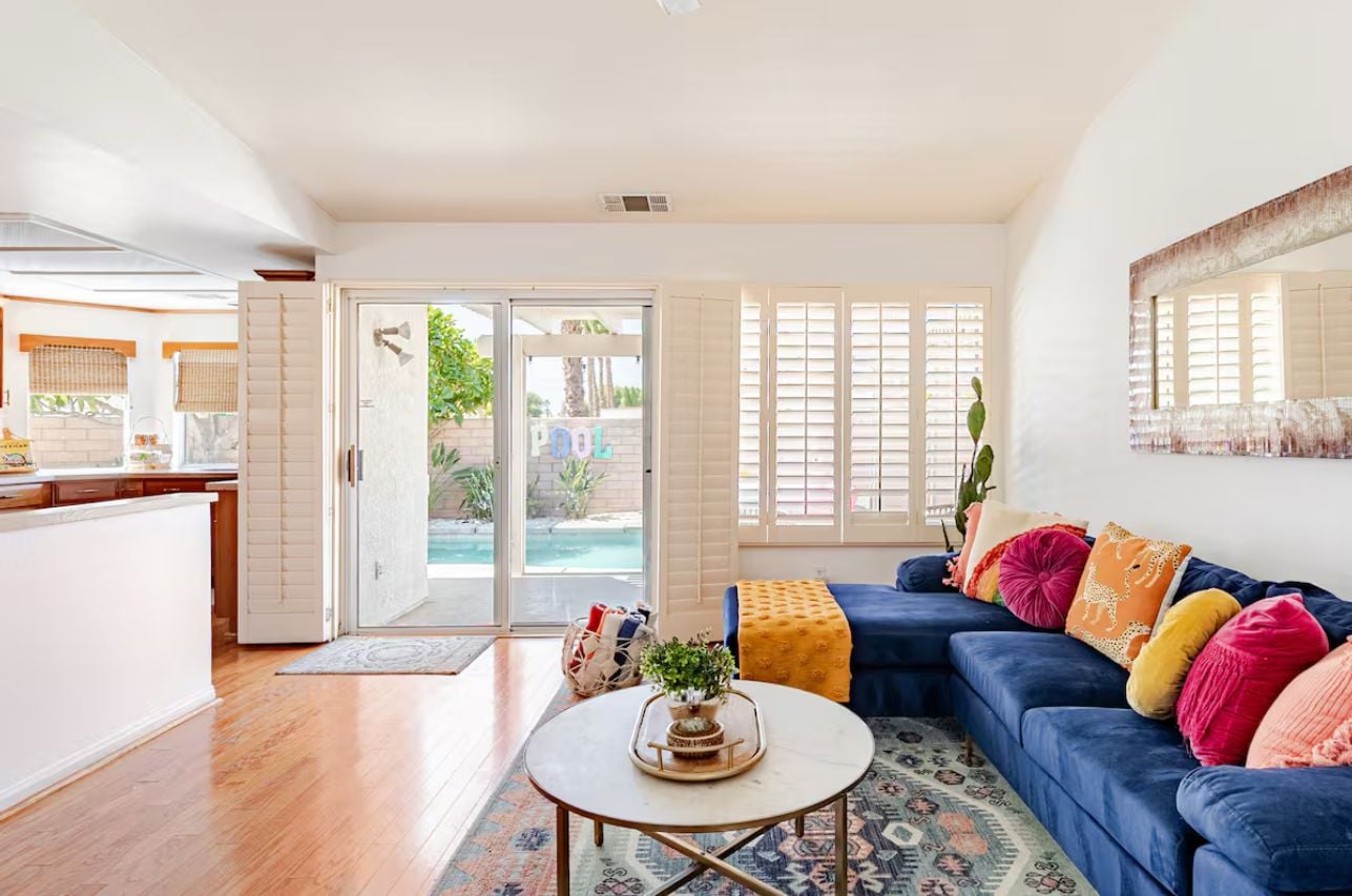 A spacious living room with a blue sofa, colorful cushions, a coffee table, and a view of a pool through glass doors.