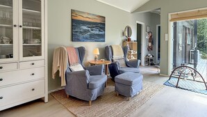 A living room with two grey armchairs, a wooden cabinet with glass doors, a painting of a seascape, and a view through a glass door to an outdoor deck. - 2 bedroom beautiful home in Gressvik (Gressvik)