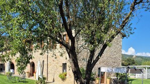 Un edificio de piedra con un gran árbol frente a él, mesas al aire libre y vistas a una colina.