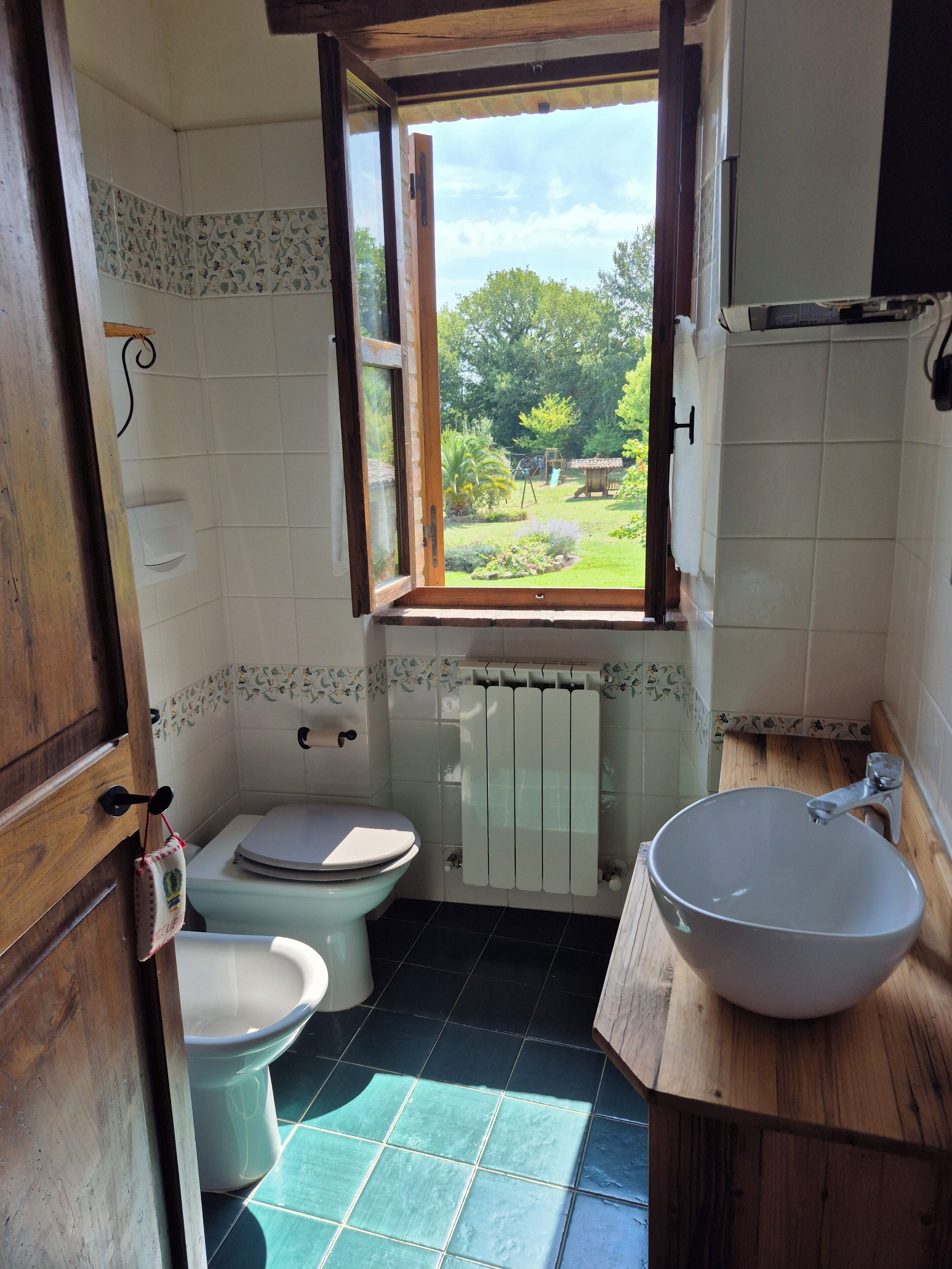 A small bathroom with a toilet, bidet, and sink. There is a window providing a view of a garden.