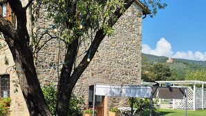 Un edificio de piedra con un patio cubierto, mobiliario exterior y un árbol en primer plano.