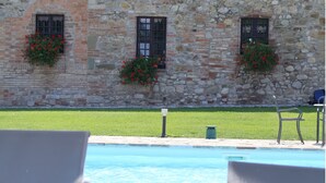 Un edificio de piedra con tres ventanas, cada una adornada con una maceta de flores. Frente a él, una piscina con sillas de descanso.