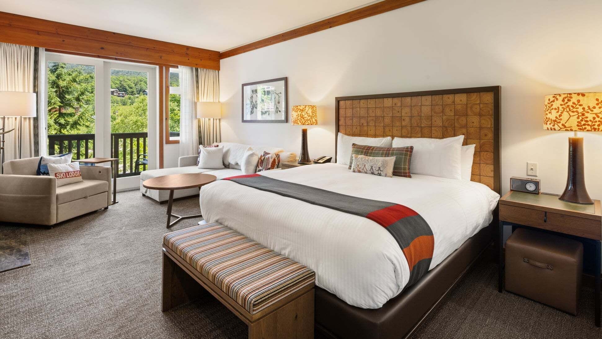 A hotel room with a large bed, a sofa, a small table, a bench, and a view of greenery through the windows.