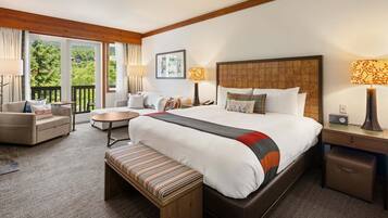 A hotel room with a large bed, a sofa, a small table, a bench, and a view of greenery through the windows.