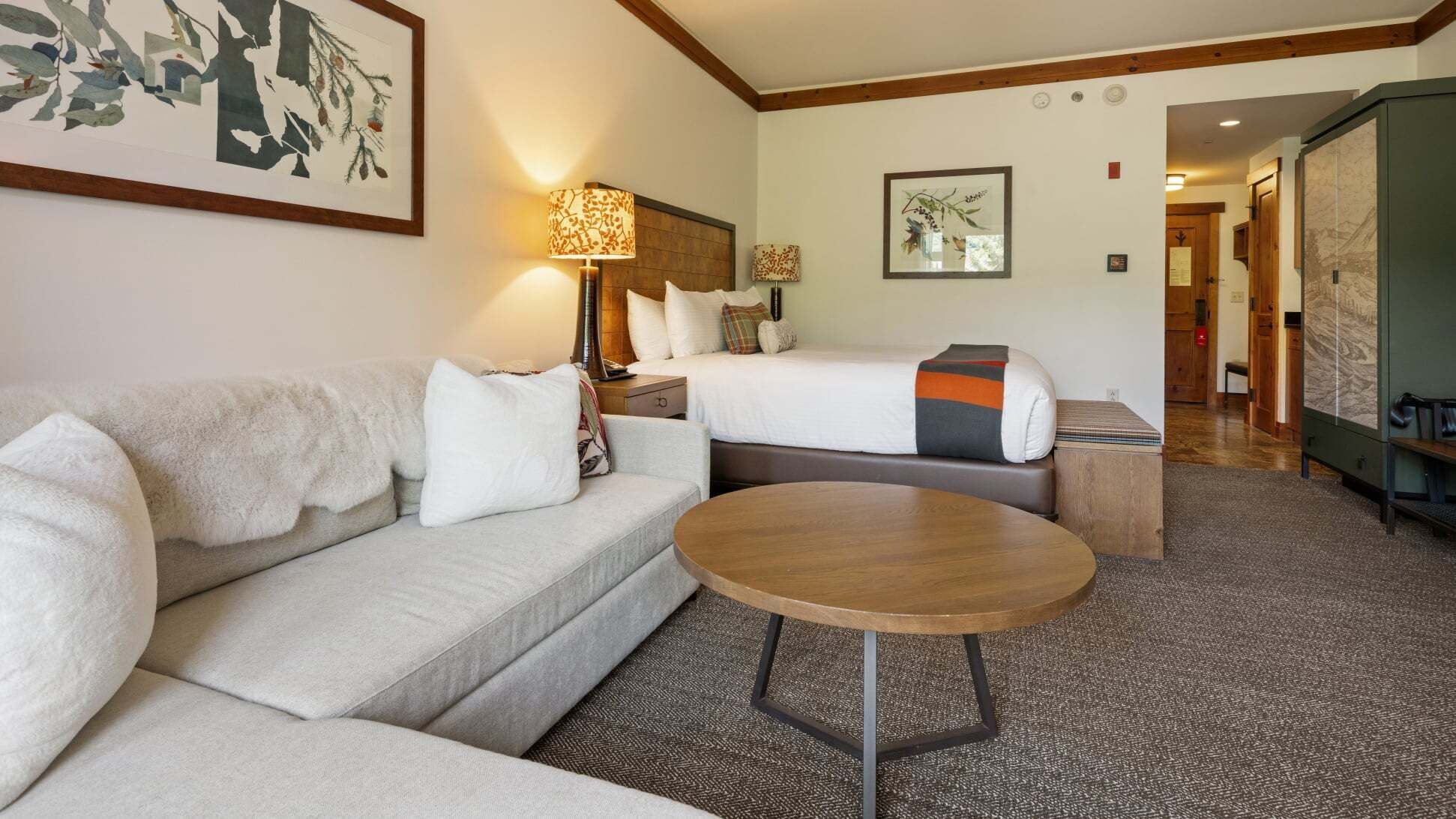 A hotel room with a sofa, a bed with a blanket, a bedside table, a lamp, and a small round table.
