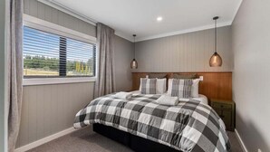 A bedroom with a large bed, a window with blinds, a nightstand, and a hanging light fixture.