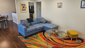 Una sala de estar con un sofá seccional, una mesa de centro de vidrio, un taburete amarillo y un tapete a rayas de colores.