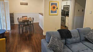 Un comedor con mesa y sillas de madera, una cocina con electrodomésticos modernos y una sala de estar con sofá y cojines.