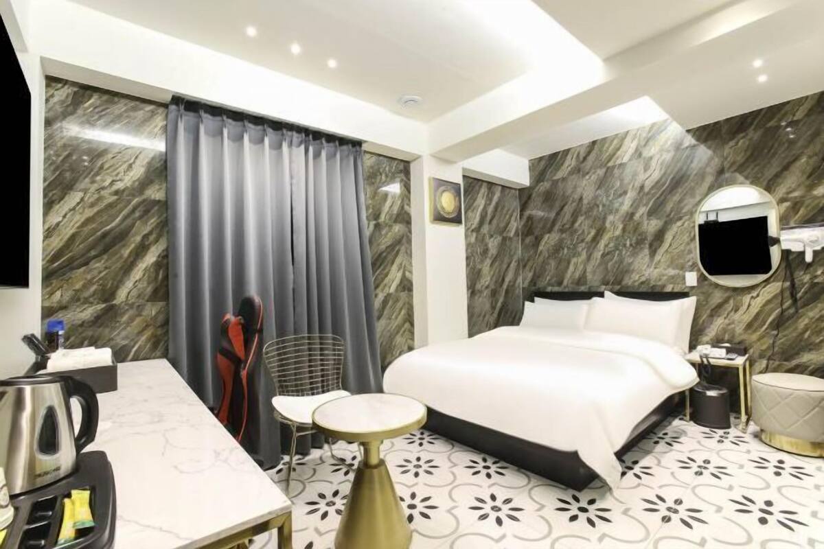 Una habitación de hotel moderna con pared de mármol, cama, escritorio con hervidor, silla y una pequeña mesa.