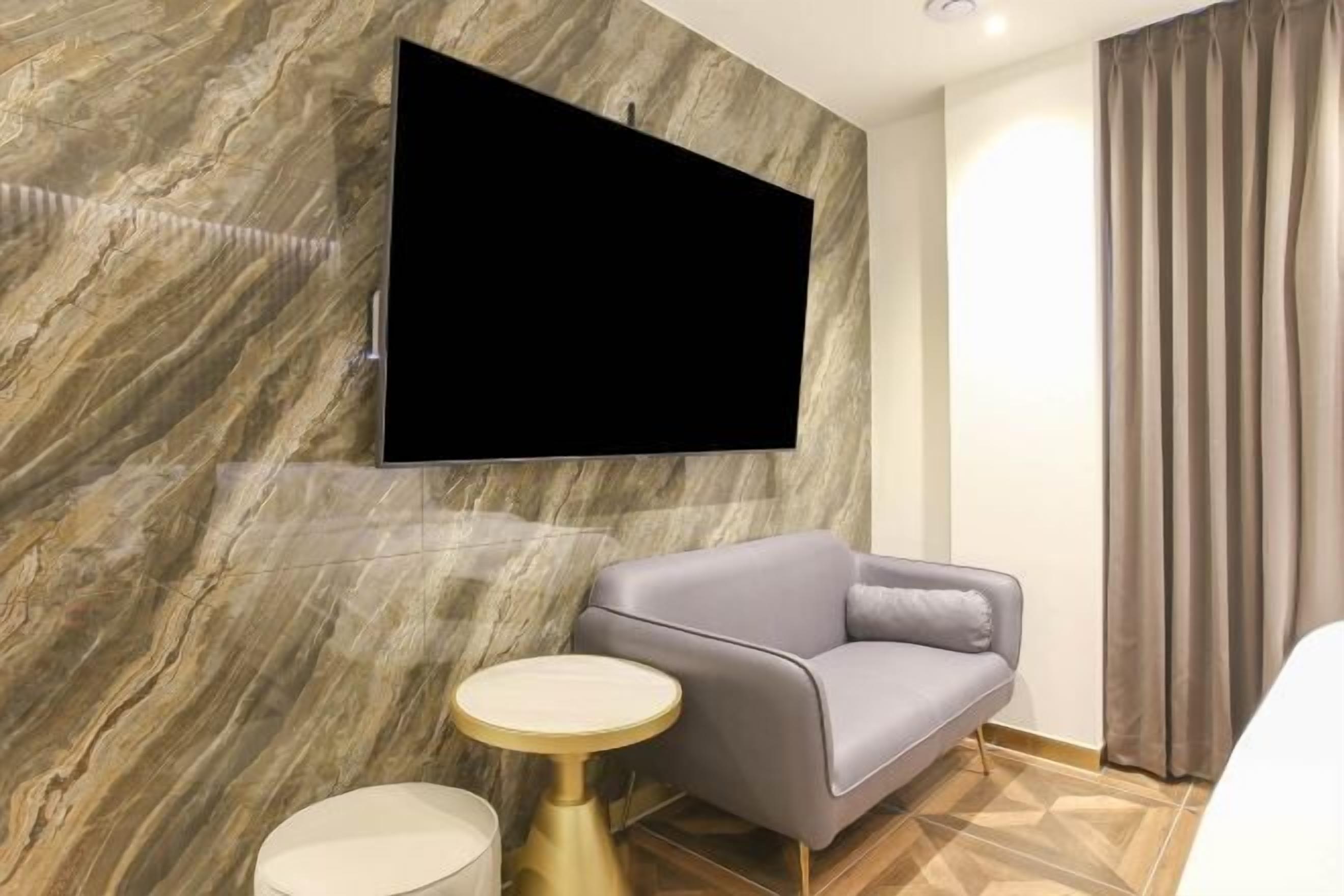 Quarto moderno com televisão na parede, sofá cinza, mesa redonda pequena e cortina.