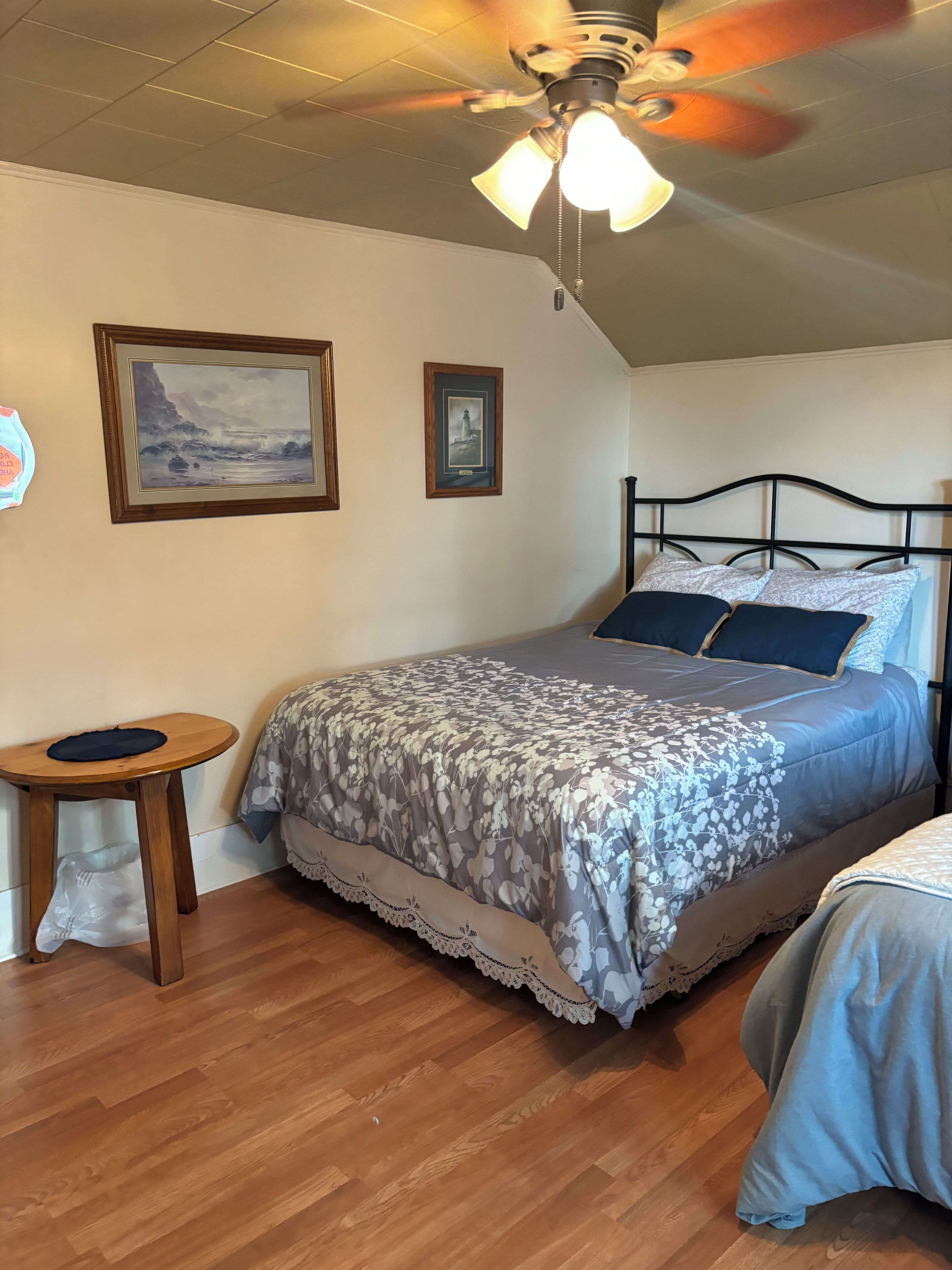 A bedroom with a bed, a ceiling fan, wooden flooring, and two framed pictures on the wall.