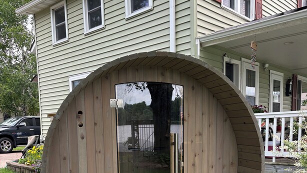 A wooden sauna structure with benches and a glass door, situated on a wooden deck with mulch, in front of a house with a porch and windows.