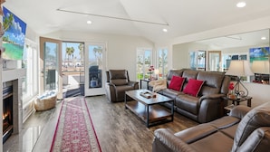 A spacious living room with a fireplace, leather sofas, a coffee table, and large windows offering a view of the outdoors. - Stunning beach home located between bay and beach w/ Ocean View Patio (San Diego)