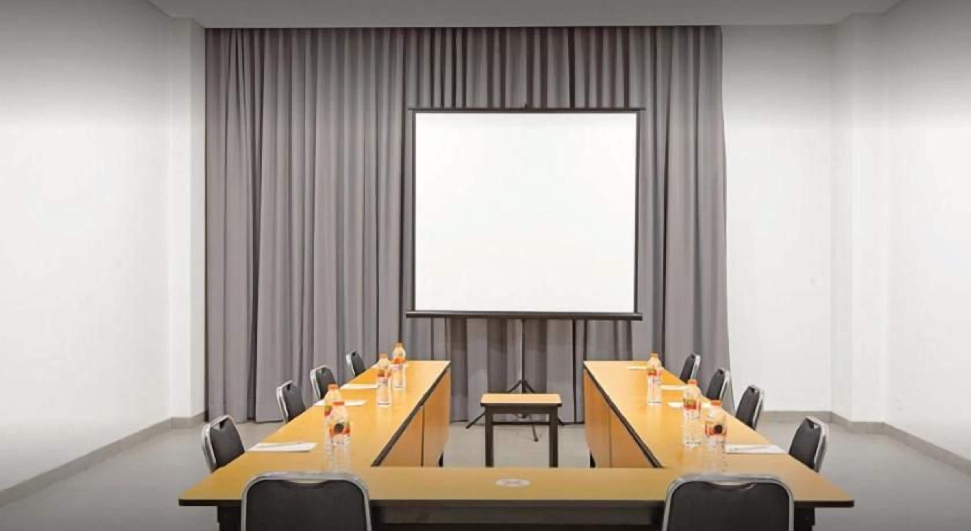 Ein Konferenzraum mit einem langen Tisch, Stühlen, einer Projektionsfläche und einer Whiteboard-Tafel.