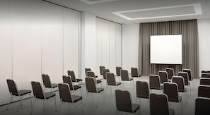 Ein Konferenzraum mit Reihen von Stühlen, die auf einen großen Bildschirm ausgerichtet sind, weißen Wänden und einer Decke mit eingelassenen Leuchten.