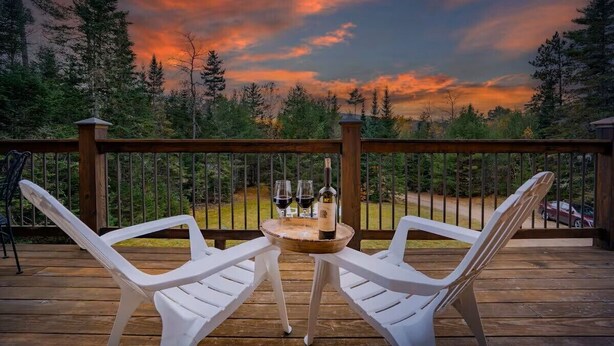 A wooden deck with two white chairs and a table, a bottle of wine, and two glasses.