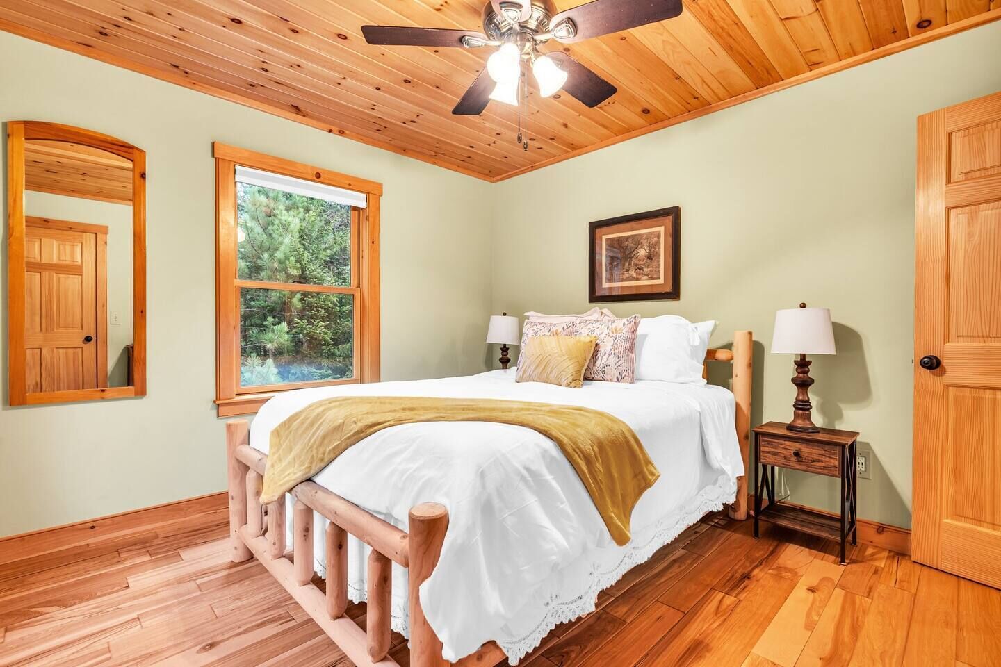 Un dormitorio con una cama de madera, un ventilador de techo, un ventanal con vistas a la vegetación y una puerta.