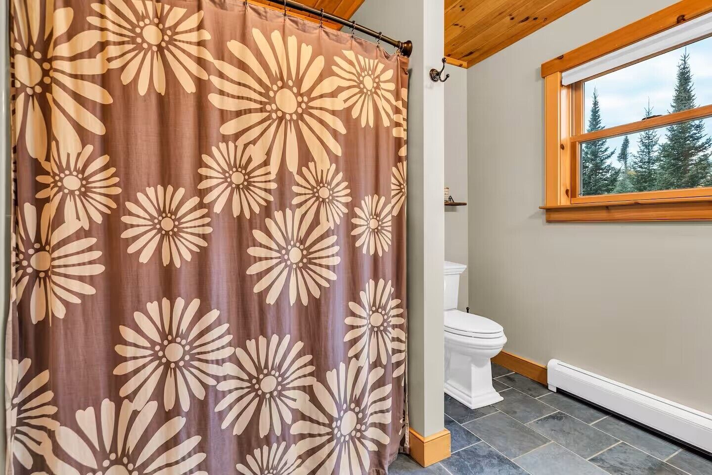Une salle de bain avec un rideau de douche orné d’un grand motif floral, des toilettes blanches et une fenêtre donnant sur des arbres.