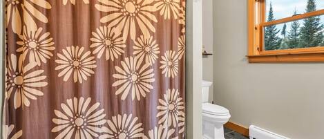Une salle de bain avec un rideau de douche orné d’un grand motif floral, des toilettes blanches et une fenêtre donnant sur des arbres.
