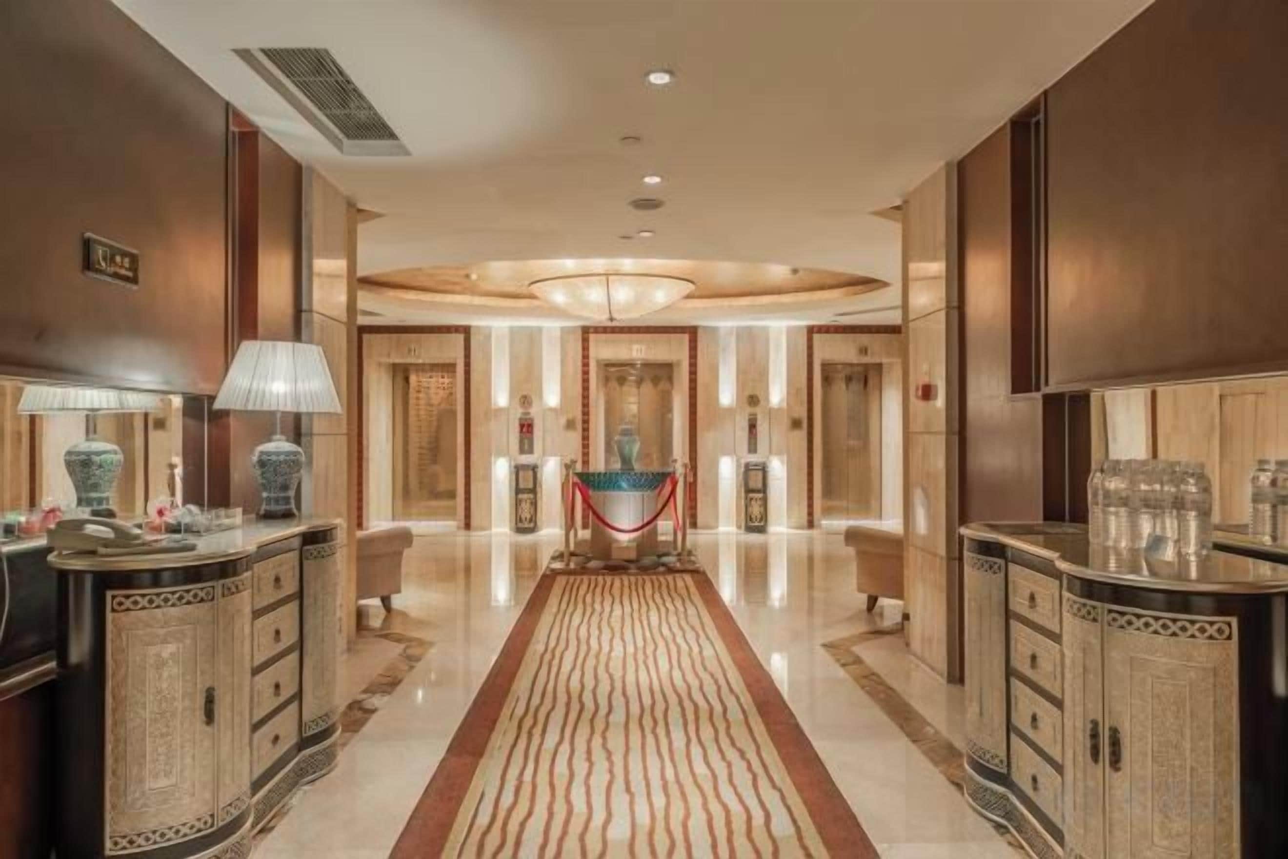 A hotel lobby with a central chandelier, a reception desk, and a red carpet leading to the entrance.