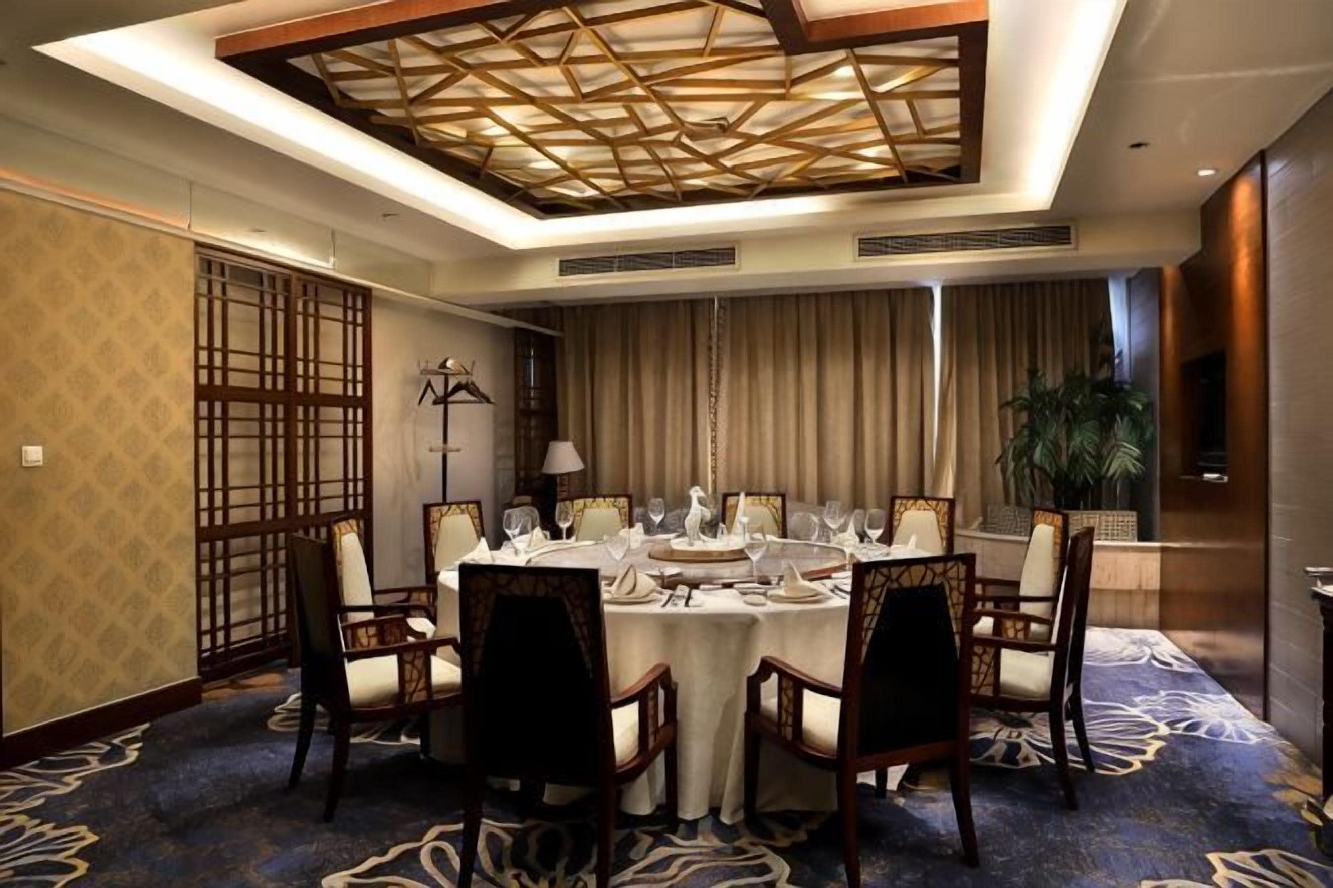 Un comedor de hotel con una mesa redonda dispuesta para seis personas, sillas alrededor, una lámpara en el techo y una planta grande.