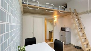 Ein kleines Zimmer mit Etagenbett, Esstisch mit Stühlen, einer kleinen Küchenzeile inklusive Mikrowelle und einer Holzleiter.
