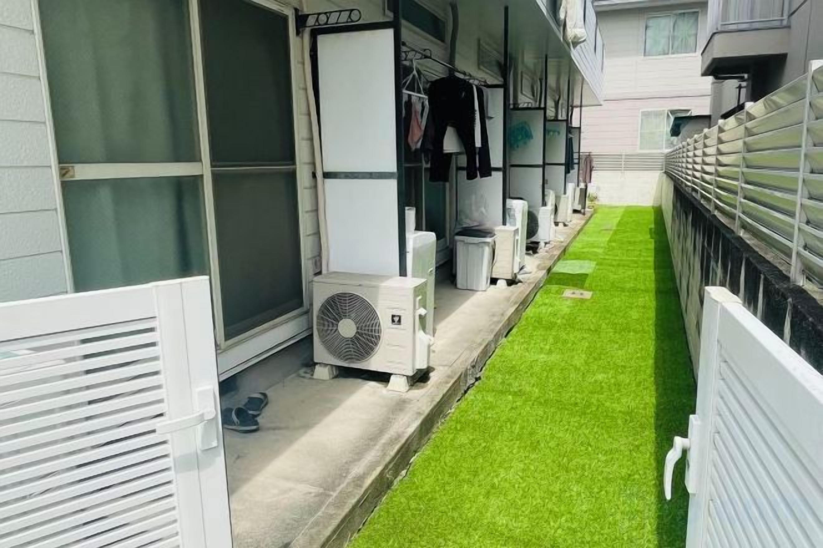 A narrow outdoor corridor with artificial grass, air conditioning units, and a white fence.