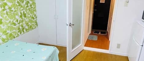 A room with a bed, a wardrobe, a door leading to a hallway, and a small refrigerator.