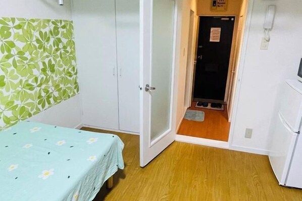 A room with a bed, a wardrobe, a door leading to a hallway, and a small refrigerator. - Fukuoka Hotel COCO (Fukuoka)