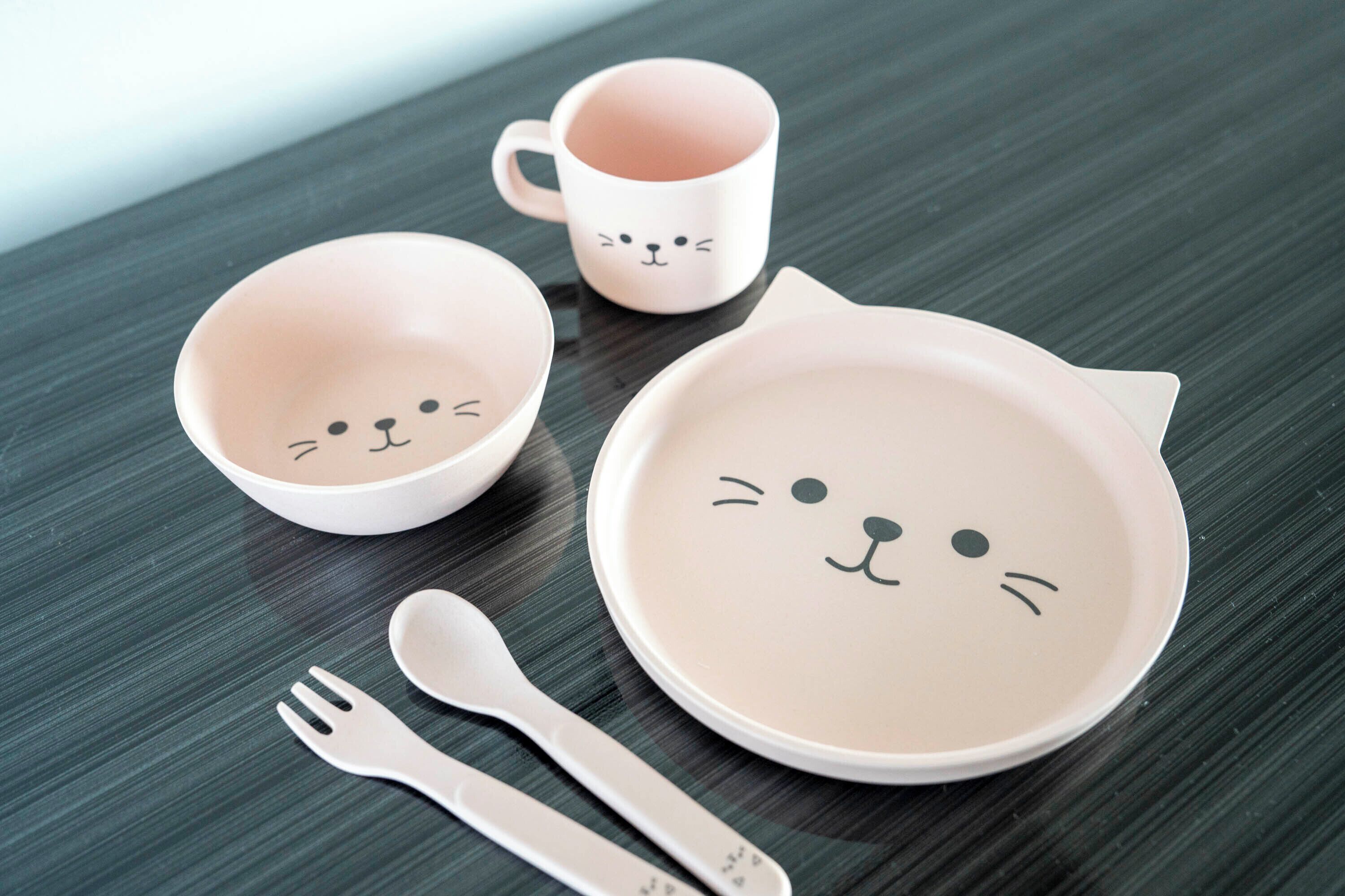 A set of white ceramic plates, bowls, and a mug with cat faces, arranged on a dark wooden surface.